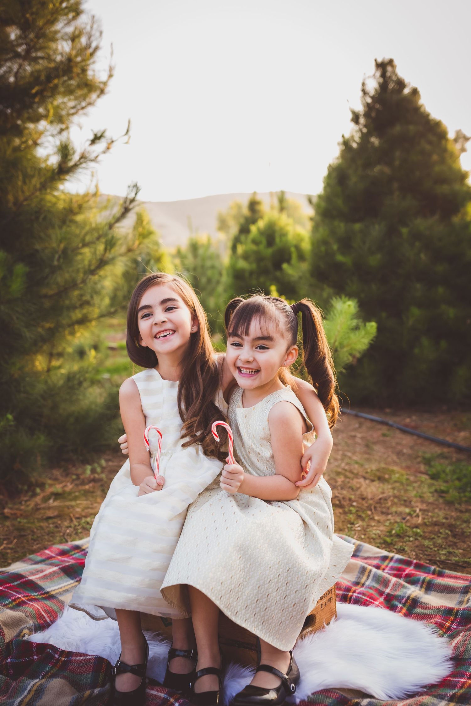 Two children in white dresses laugh together while sitting on a plaid blanket outdoors with evergreen trees in background.