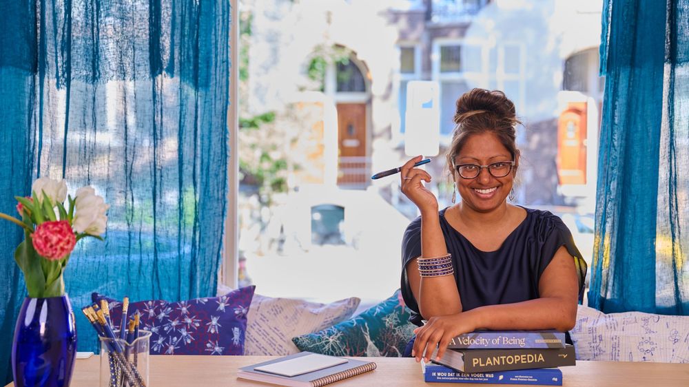 A person wearing glasses sits at a desk with blue curtains and a vase of flowers in a bright sunlit room.