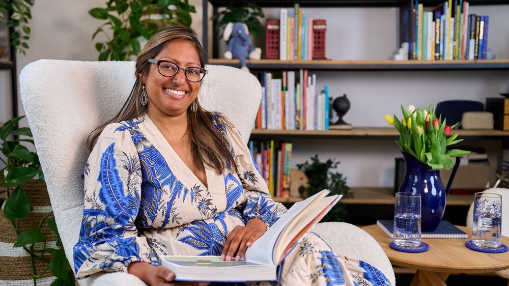 Cheerful person wearing glasses and a blue and white floral top writes at a desk with bookshelves in the background.