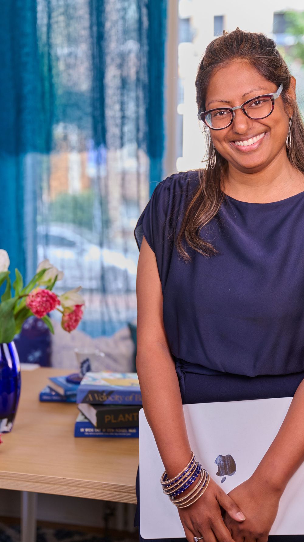 Professional wearing glasses and a navy blouse while smiling at their desk in an office setting.