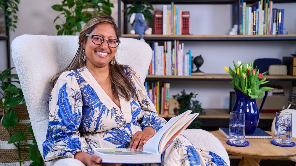 A person in a blue patterned robe sits in a white armchair with bookshelves and plants in the background.