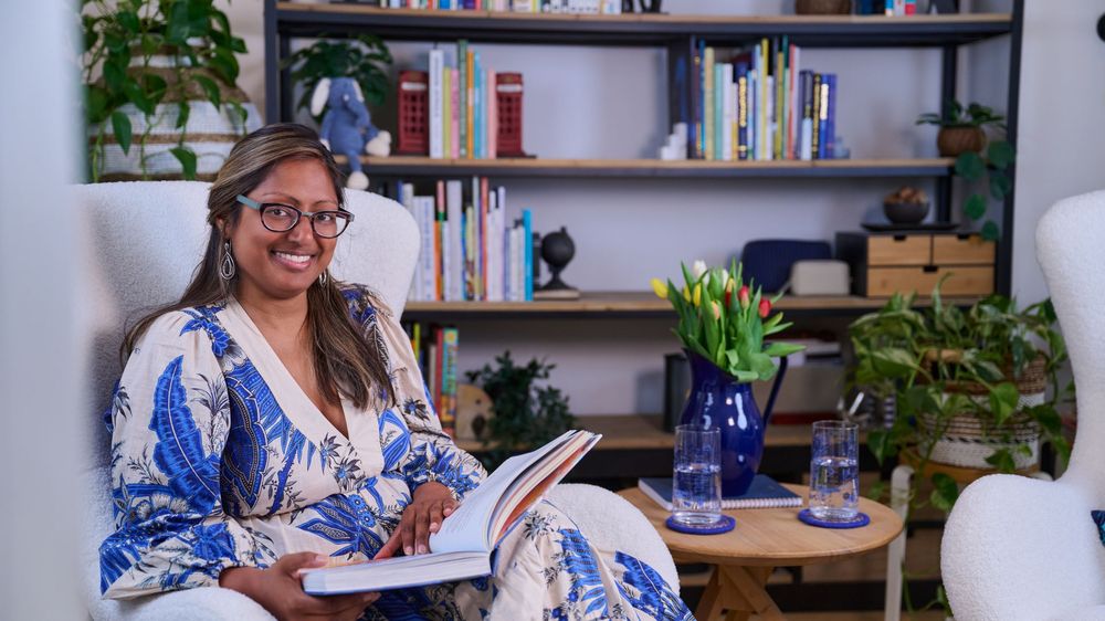 A person sits at desk writing in cheerful, well-lit room with filled bookshelves and plants in the background.