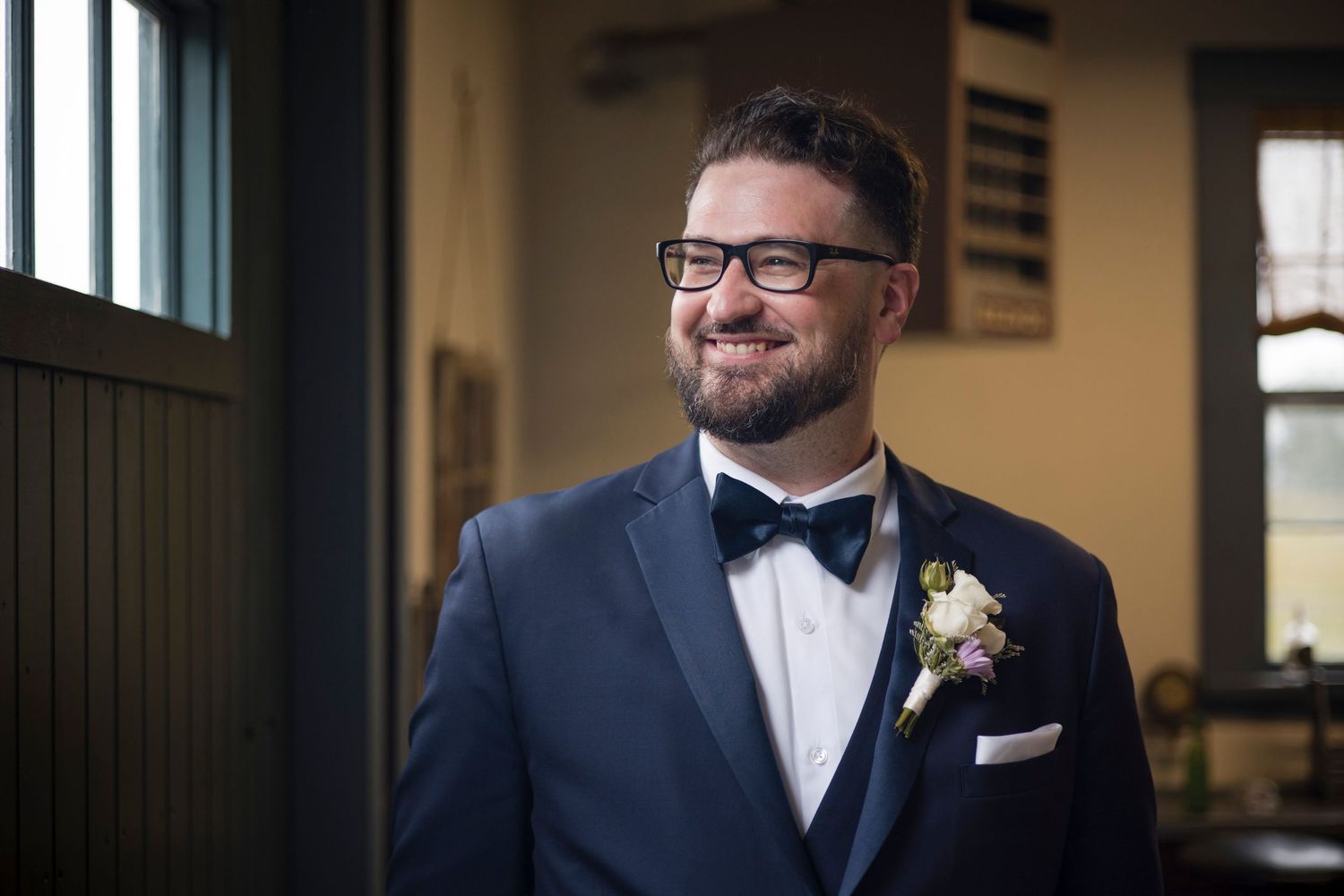 Smiling groom in a navy suit and bow tie wearing glasses with a boutonniere at a wedding venue.