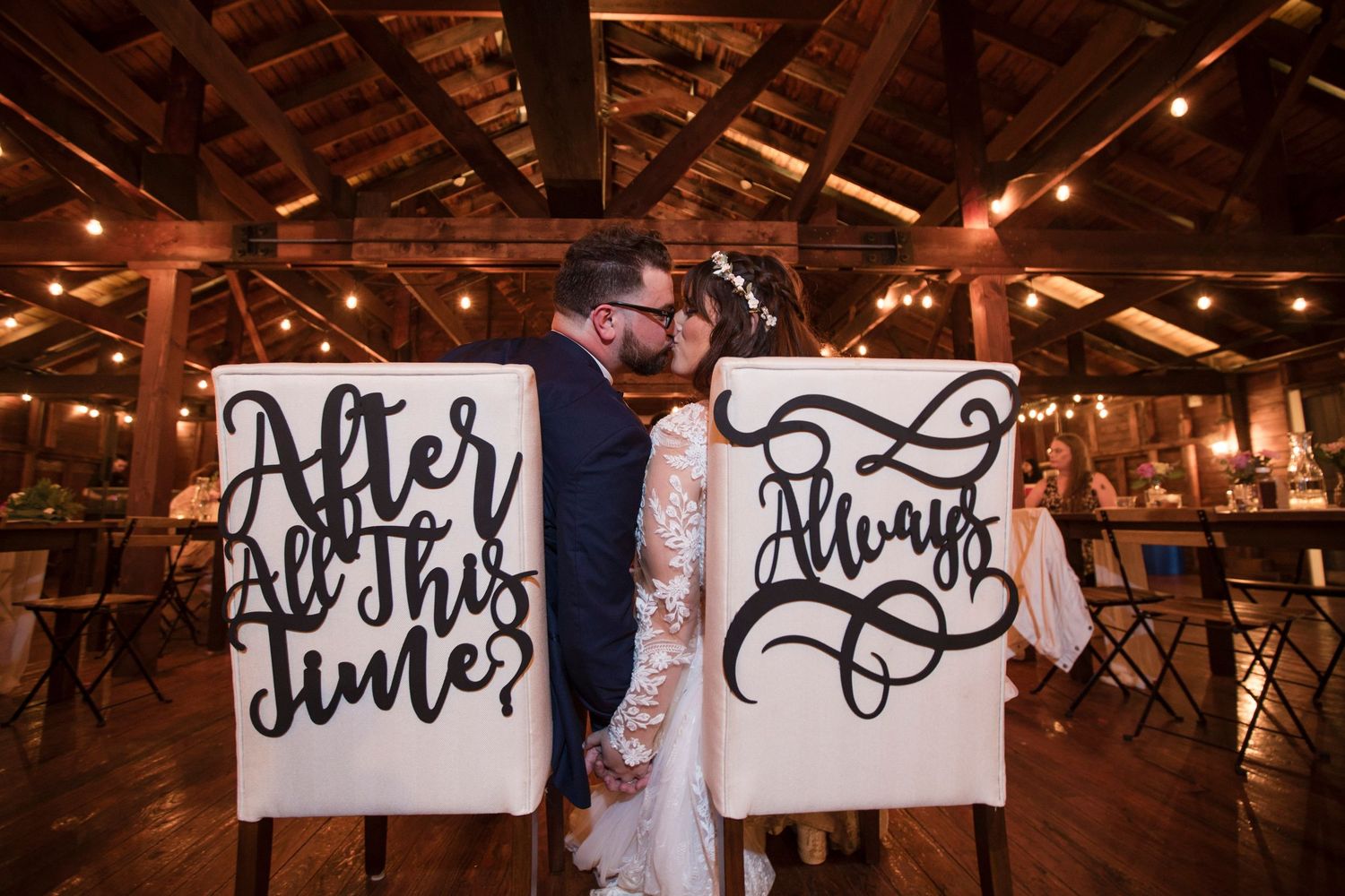 A rustic barn wedding reception with two decorative signs reading 'After All This Time' and 'Always' displayed on chairs.