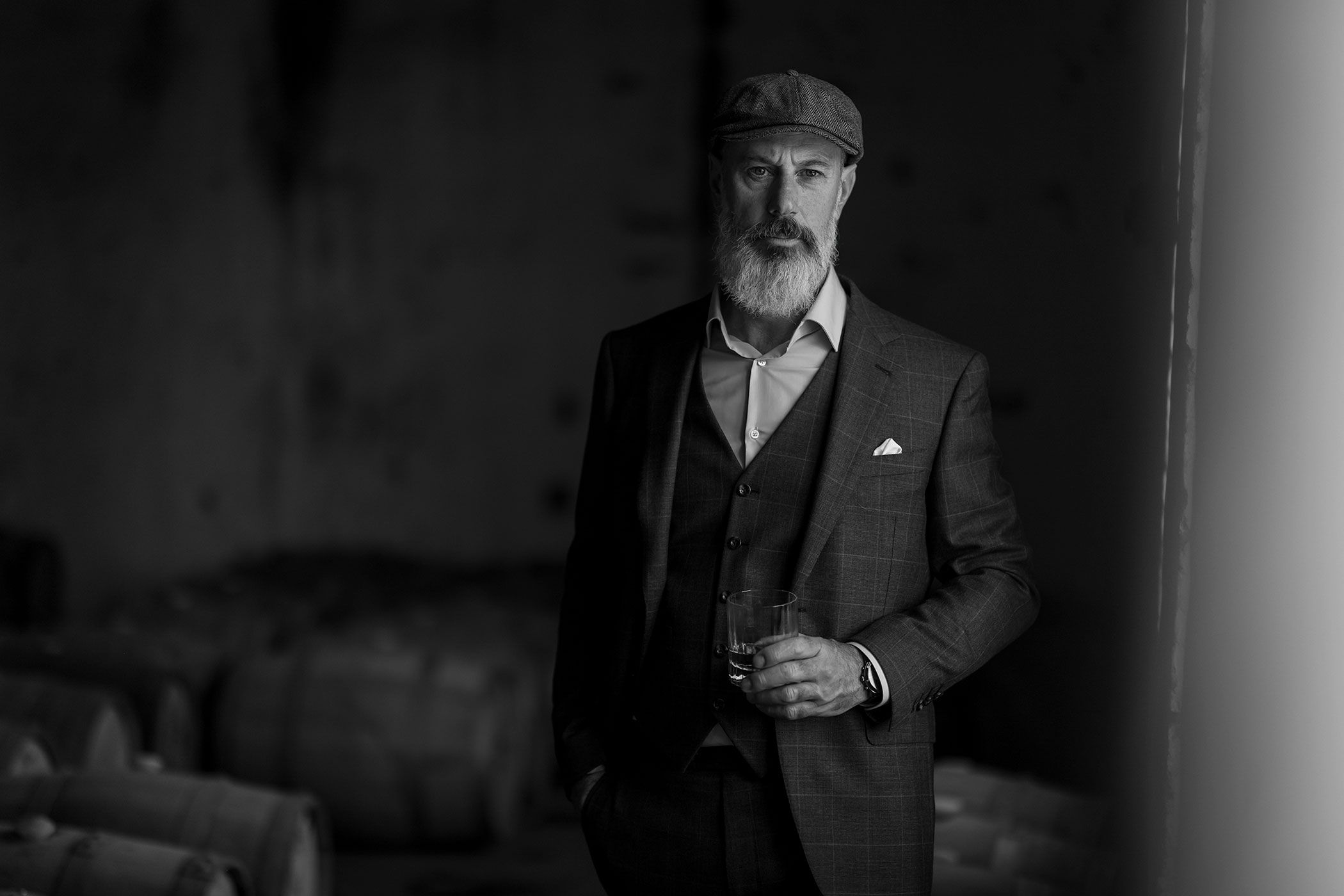 Black and white portrait of gentleman in vintage attire with flatcap and three piece suit standing indoors.