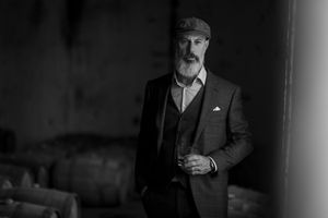 Black and white portrait of gentleman in vintage attire with flatcap and three piece suit standing indoors.