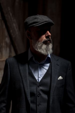 Dramatic portrait of a bearded man in a dark suit, vest and flat cap gazing to the side in moody lighting.