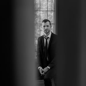 Man in a black suit stands in dark dramatic lighting near a window creating a moody film noir aesthetic.