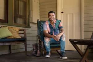 Guy Sebastian sits in a casual pose on a turquoise chair on a rustic wooden porch with boots nearby.