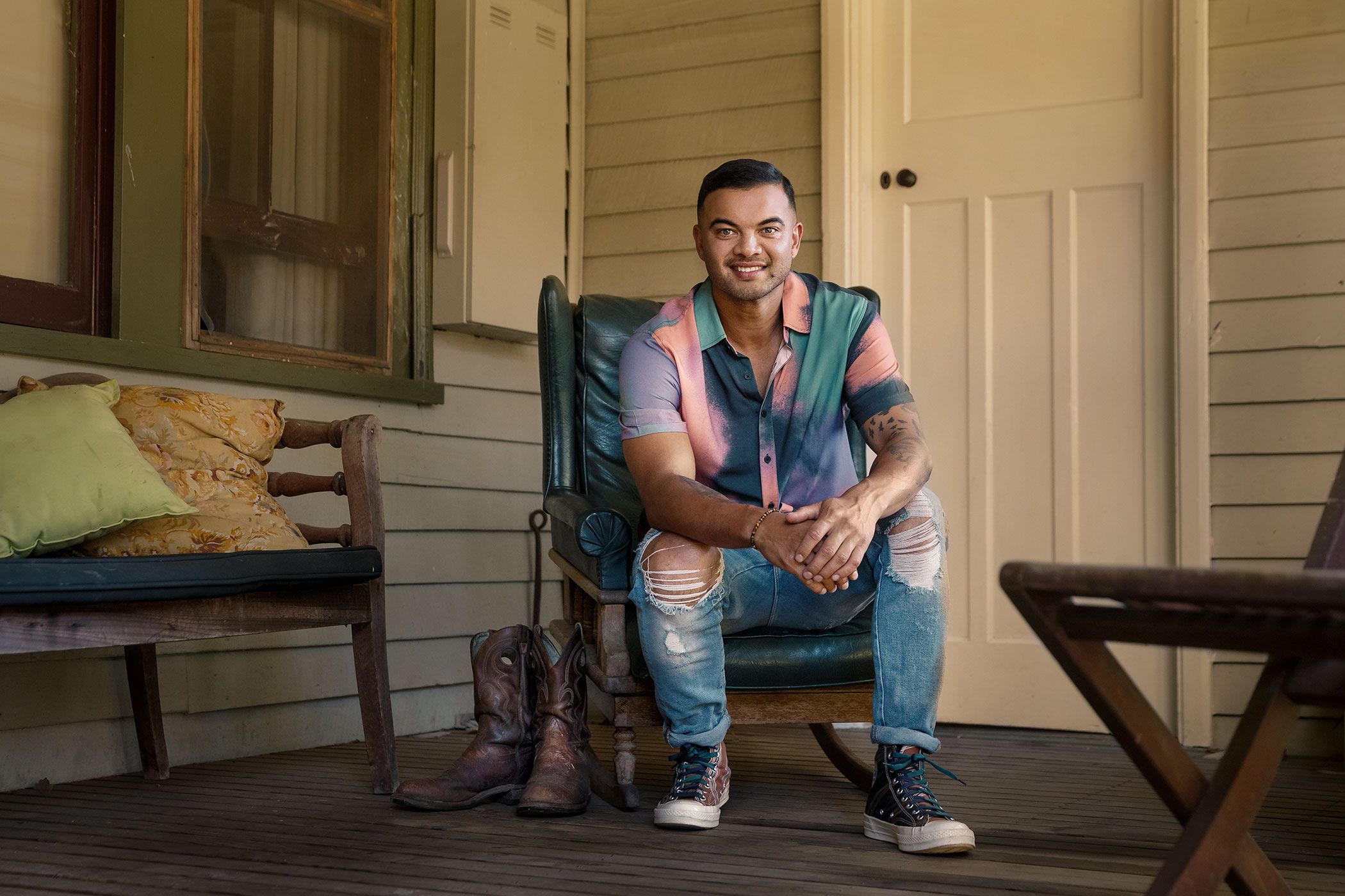 Guy Sebastian sits in a casual pose on a turquoise chair on a rustic wooden porch with boots nearby.