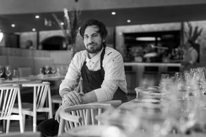 A restaurant Head Chef in an apron sits thoughtfully at an empty table in a black and white dining room interior.