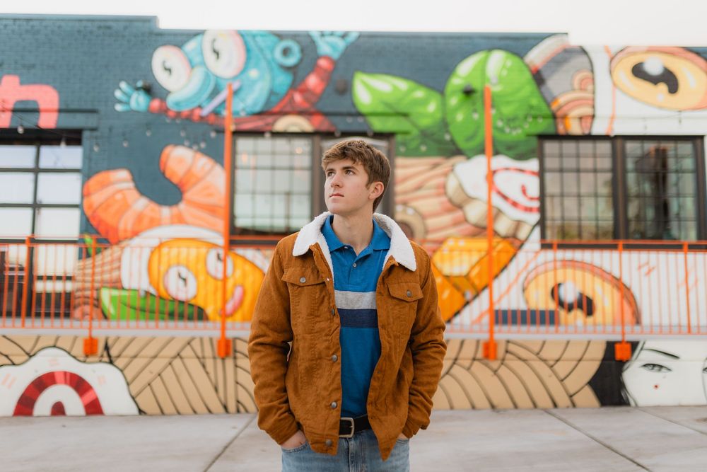 Person in brown jacket stands against colorful graffiti wall with cartoon-style street art in urban setting.