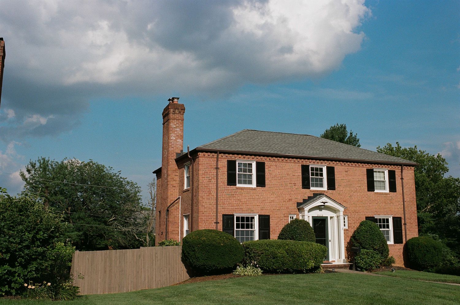 A classic brick colonial style house with black shutters under a partly cloudy sky.