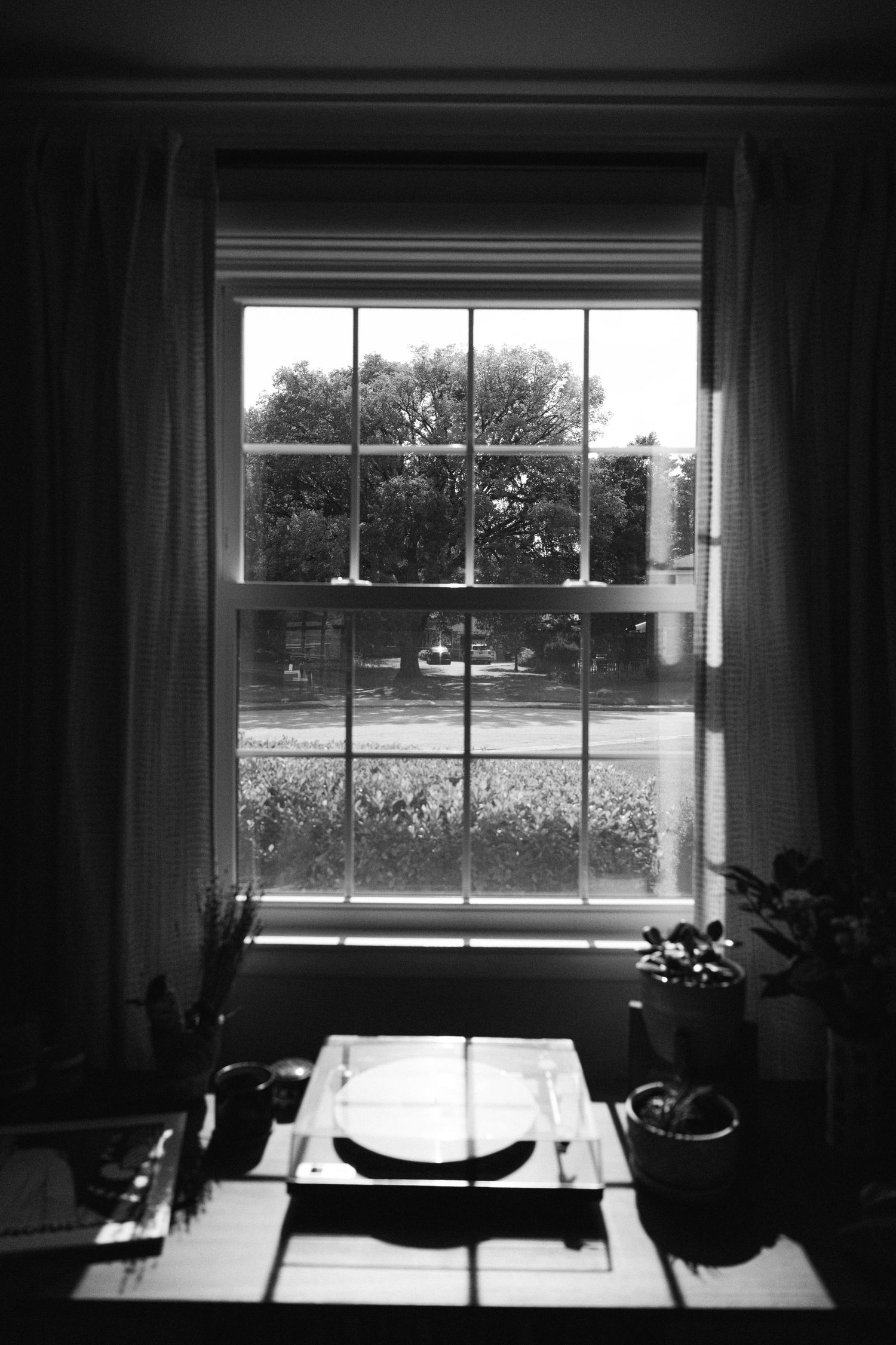 Black and white photo of a desk by a window with curtains, showing a place setting and potted plant silhouetted against daylight.