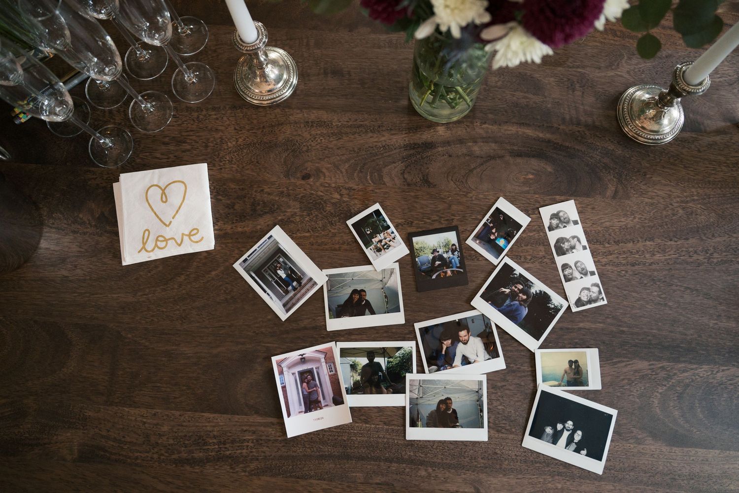 Scattered polaroid photos and heart decoration on dark wooden table with wine glasses and flowers.