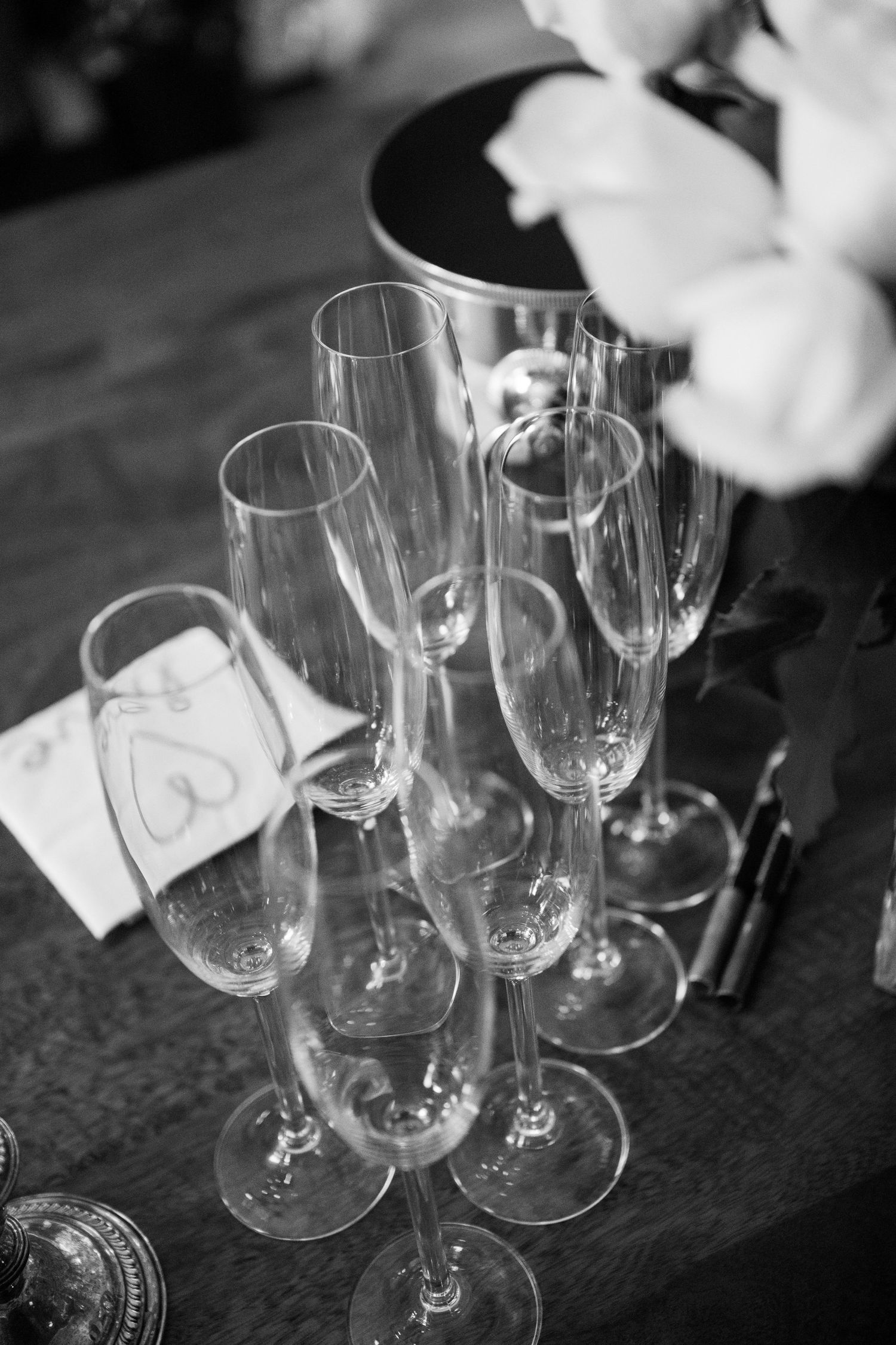 Empty champagne flutes arranged on a dark surface in black and white with soft focus florals in background.