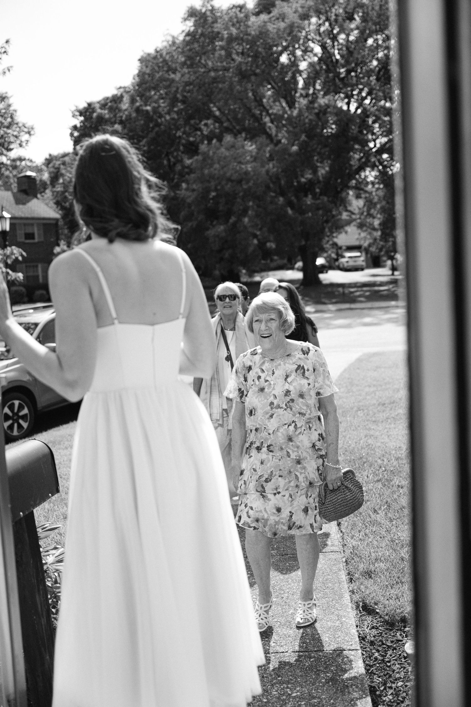 A black and white photo showing someone in a white wedding dress greeting guests outside on a sunny day.