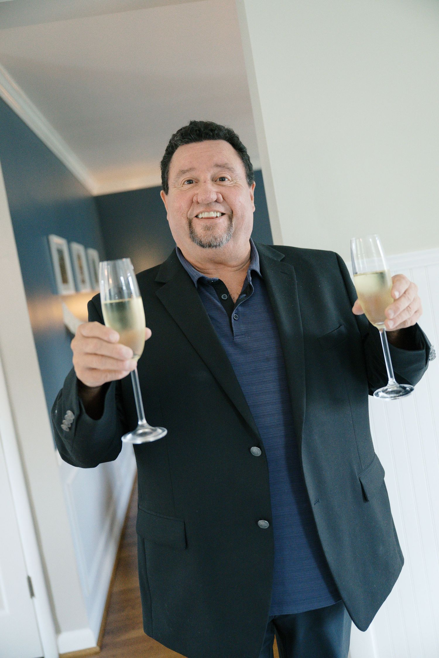 Someone in a dark blazer and navy shirt holding up two champagne glasses with a celebratory expression.