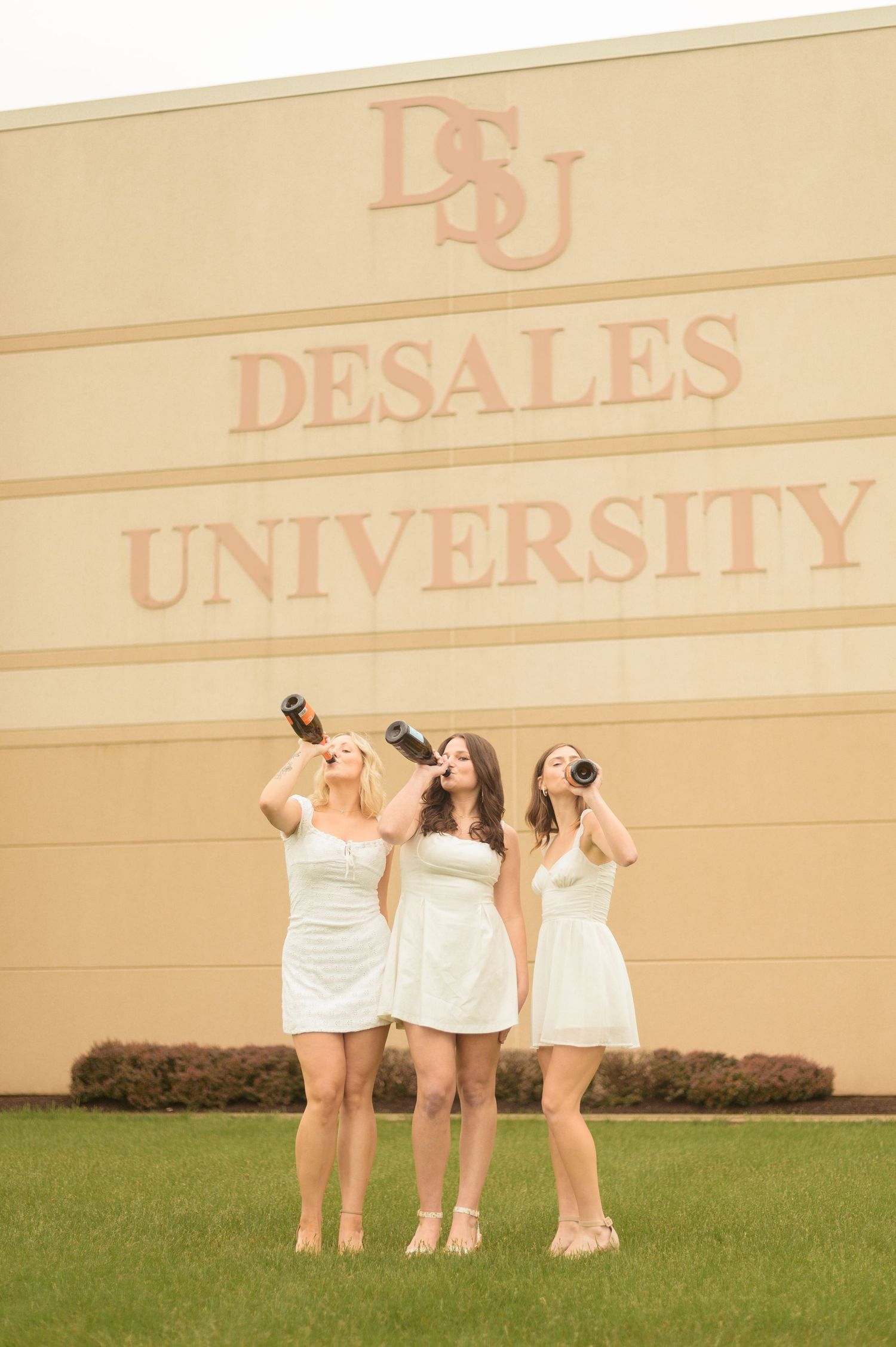 DeSales graduates in white graduation dresses casually on grass during spring season.
