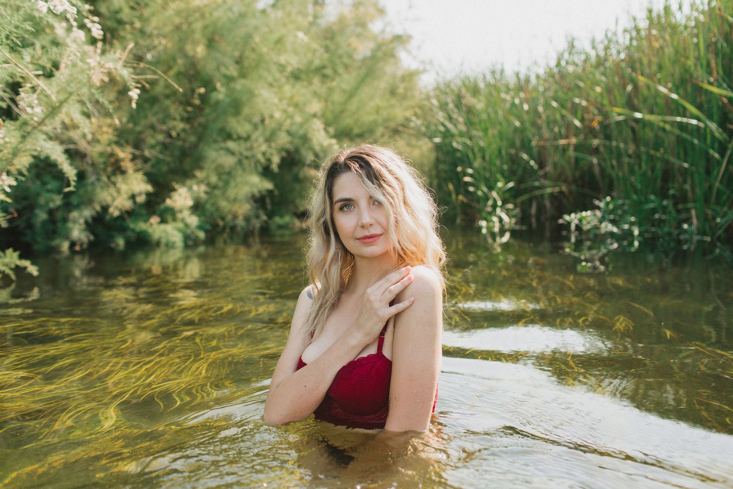 Person in burgundy swimsuit stands in shallow water surrounded by greenery on sunny day.
