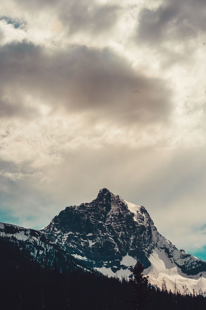 Dramatic mountain peak with snow covered slopes rises beneath moody cloudy skies in a scenic landscape view.
