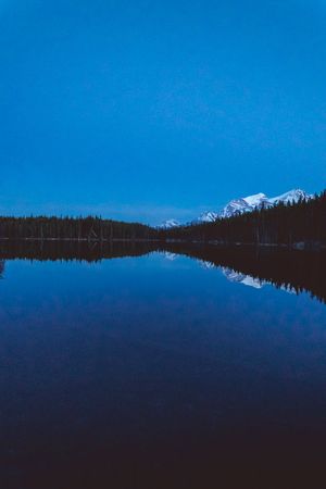 Serene mountain lake reflects snow-capped peaks and dark pine forest under a deep blue twilight sky.