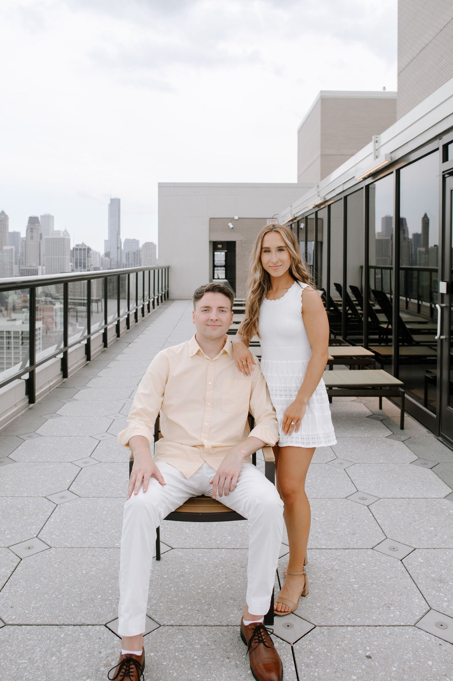 A couple in light colored outfits poses on a modern rooftop terrace with city skyline views in the background.