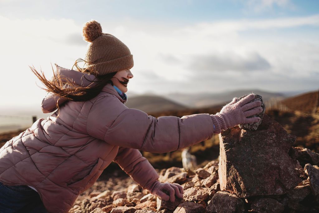 Person in pink winter coat reaches out while standing on rocky terrain during golden hour.