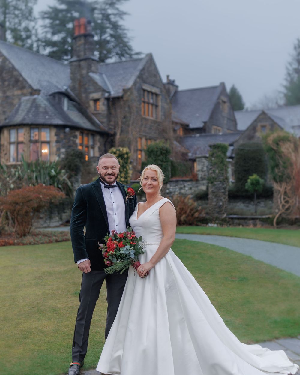 Wedding couple shares romantic winter moment on landscaped lawn of historic stone manor buildings.