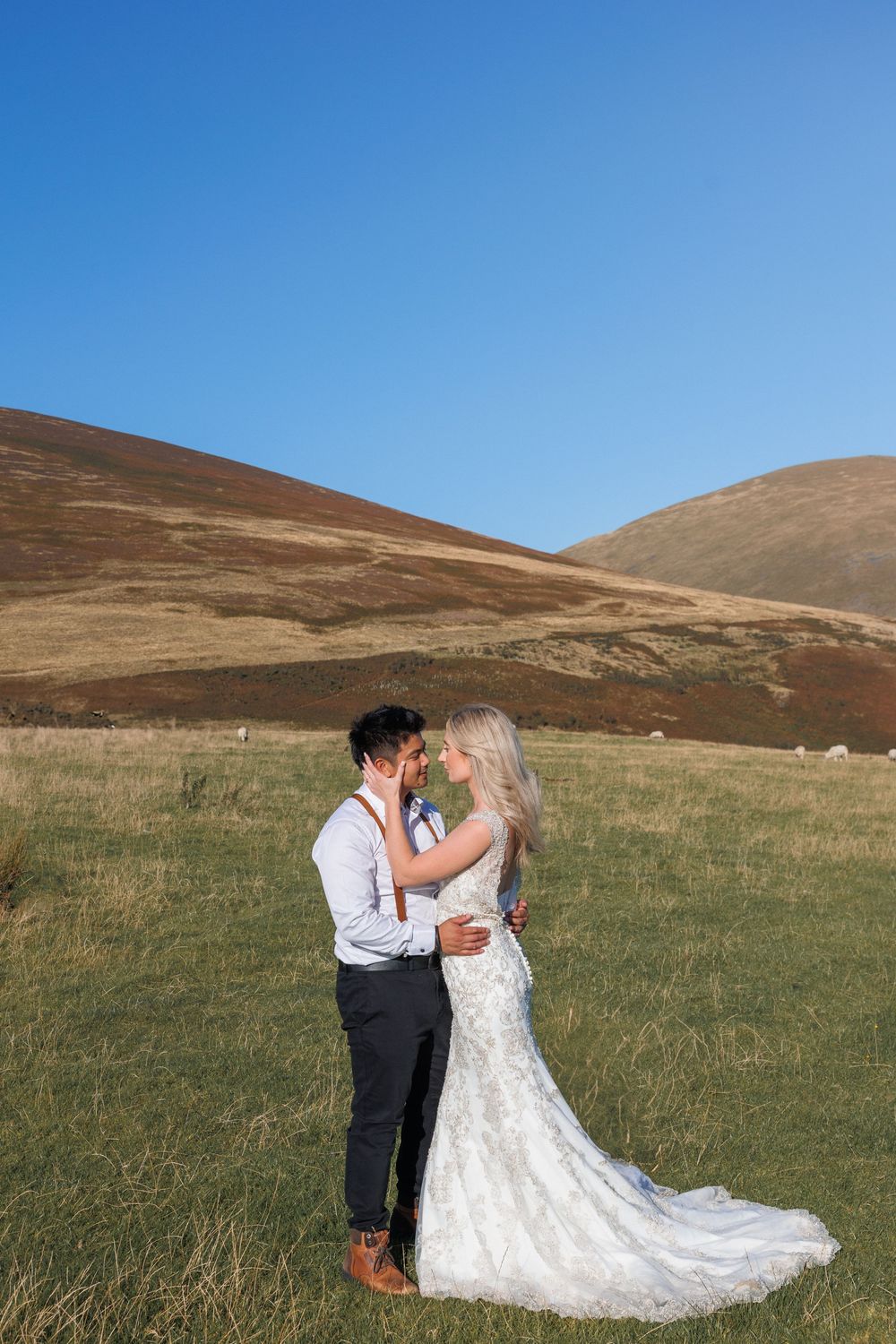 A wedding couple embraces on a grassy hillside with brown mountains and bright blue sky in the background.