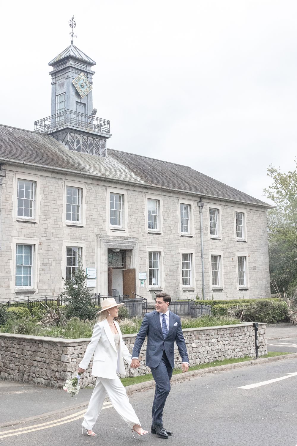A couple in wedding attire walks outside a historic stone building in characterful gray stone architecture.