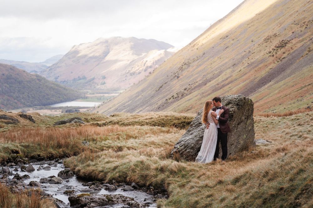 Two people stand together in the rolling hills and mountains of what appears to be the Scottish Highlands near a flowing stream.