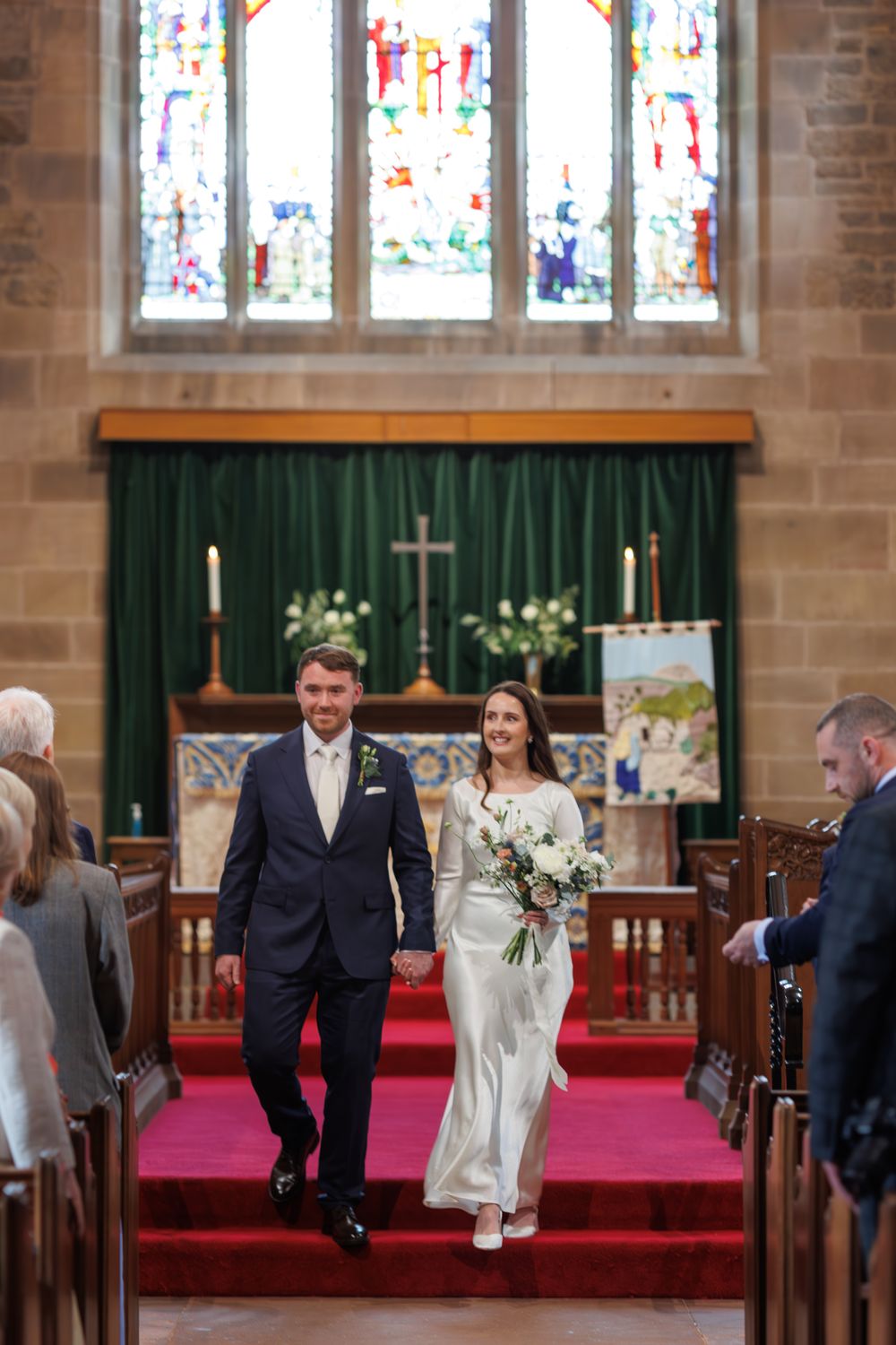 A wedding ceremony in a church with stained glass windows, green altar draping, and red carpet in the aisle.