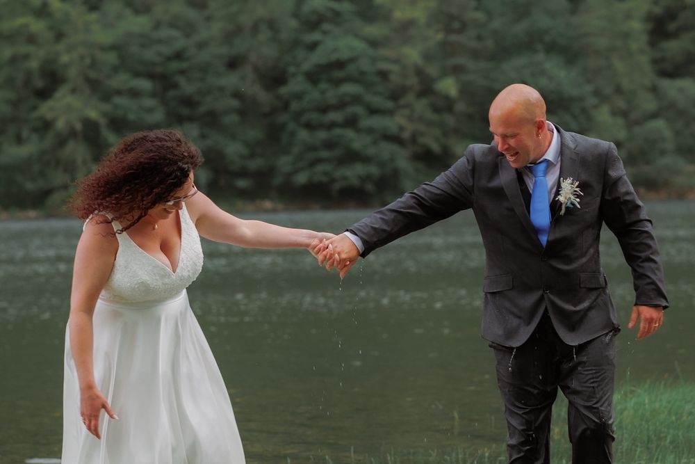 A wedding couple holds hands playfully on a lakeshore with evergreen trees in the background.