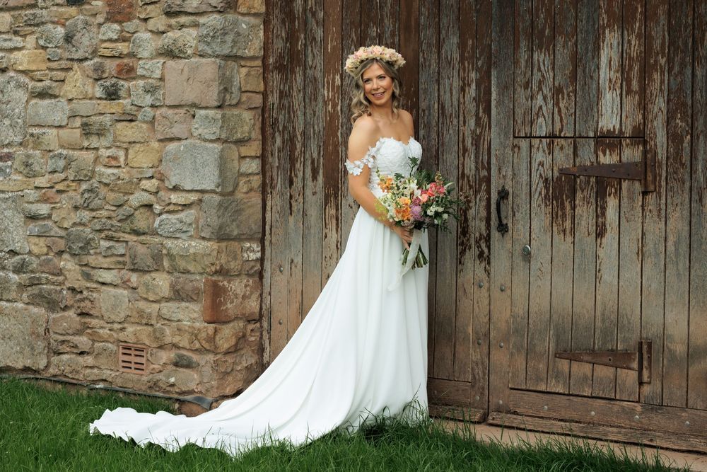 A wedding photoshoot outside a rustic stone wall and wooden door featuring a white bridal gown and orange bouquet.