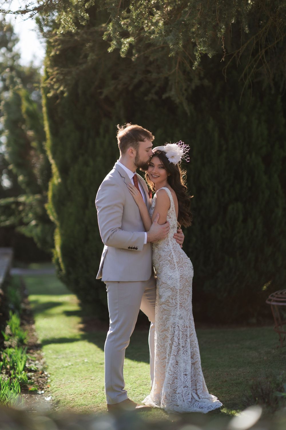 Newlyweds share an intimate moment in a garden setting with sunlight filtering through cypress trees.