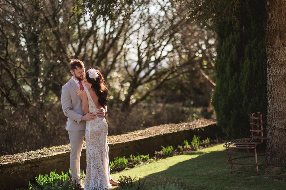 A couple in wedding attire embraces outdoors during golden hour sunlight in a garden setting.