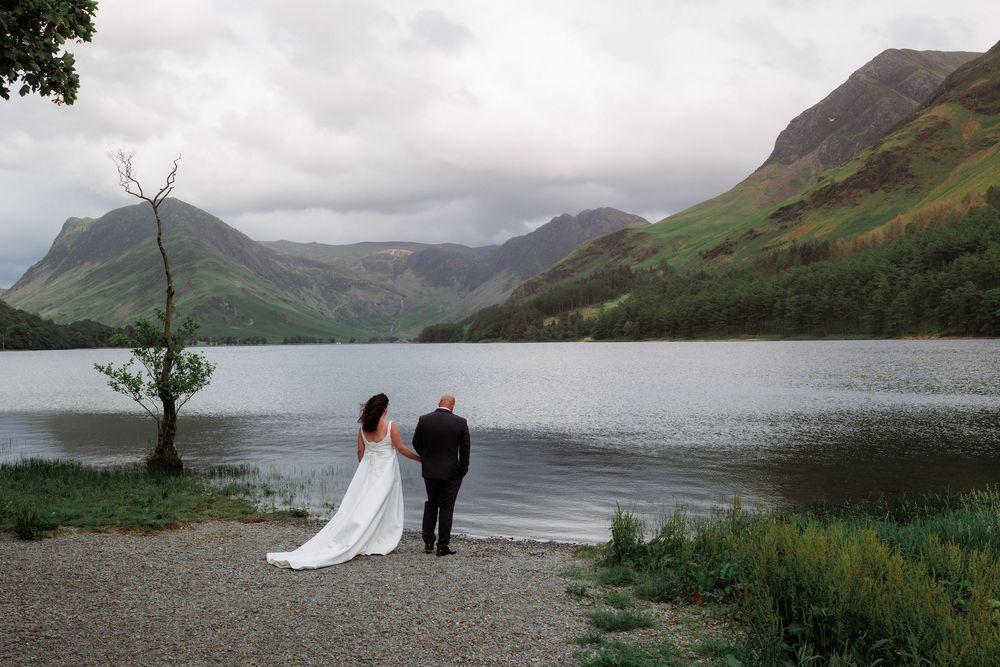 A wedding couple stands by a scenic mountain lake surrounded by green hills on a cloudy day.