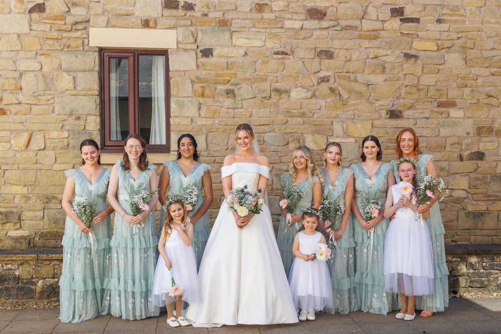 A bridal party in matching mint green bridesmaid dresses and white flower girl dresses stands against a stone wall.