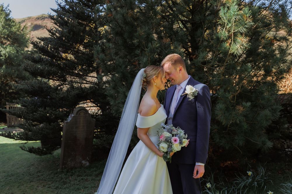 A couple in wedding attire shares a tender moment in front of evergreen trees while holding a floral bouquet.