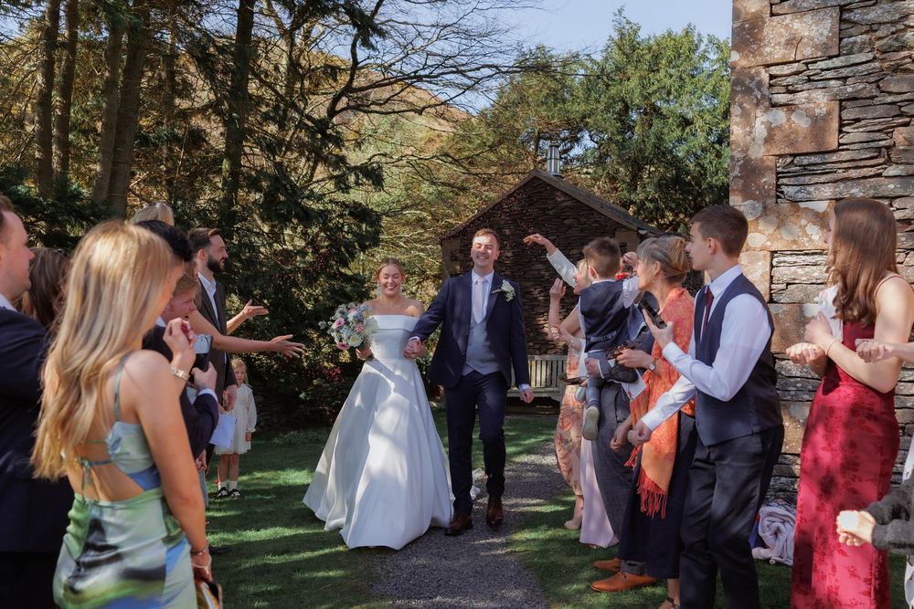 Wedding celebration with guests gathered outdoors near stone building with trees and greenery in the background.