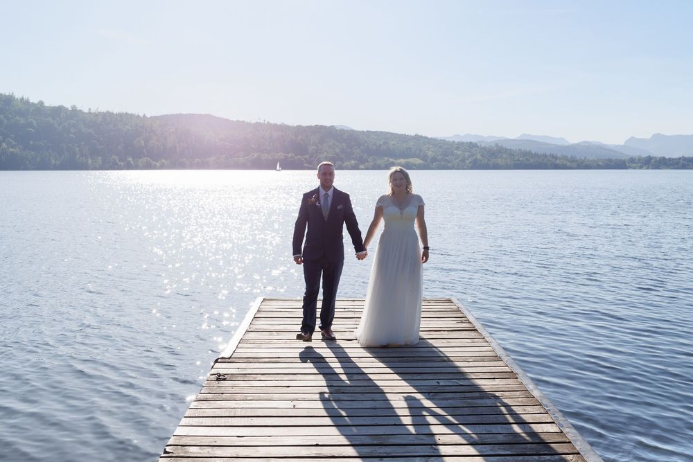 A couple in wedding attire walks hand in hand down a wooden dock over a serene mountain lake on a sunny day.