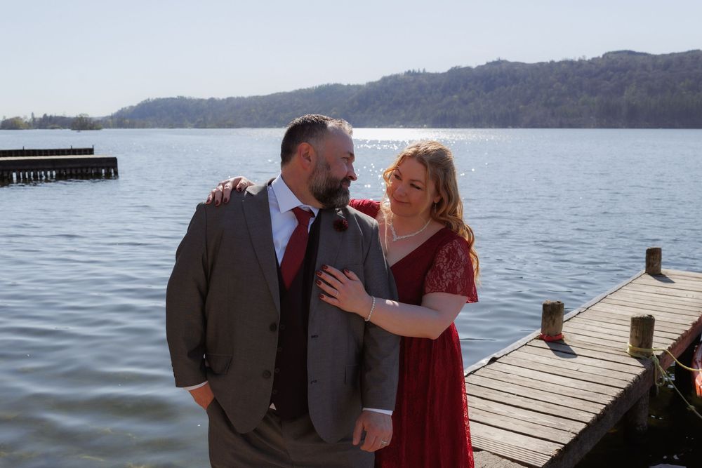 A romantic couple in formal attire share a joyful moment together on a wooden dock overlooking a serene lake.