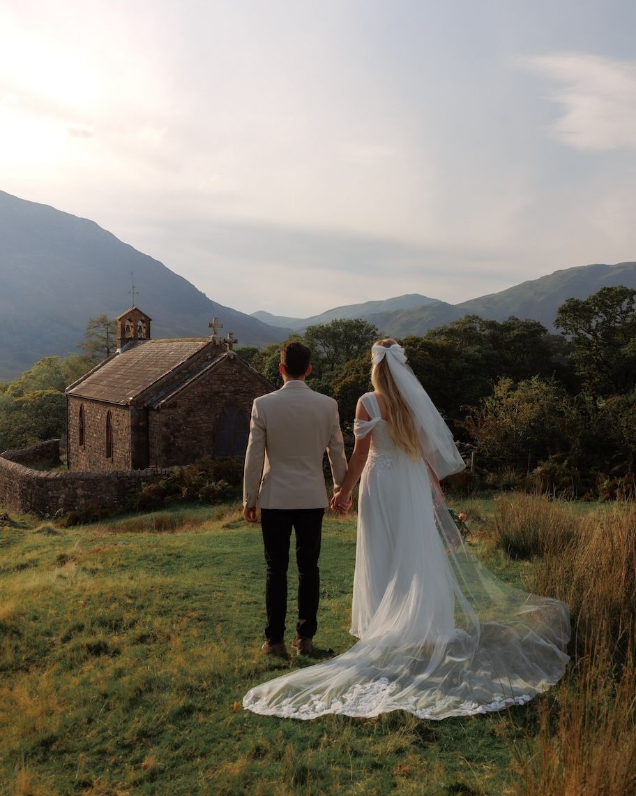 A couple in wedding attire walks away into a scenic mountainous landscape with an old stone church in the distance.