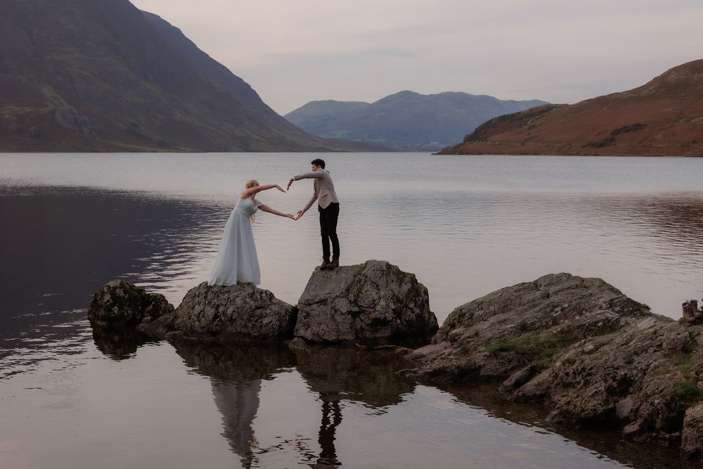 Two figures stand on rocks by a misty lake surrounded by mountains during golden hour.