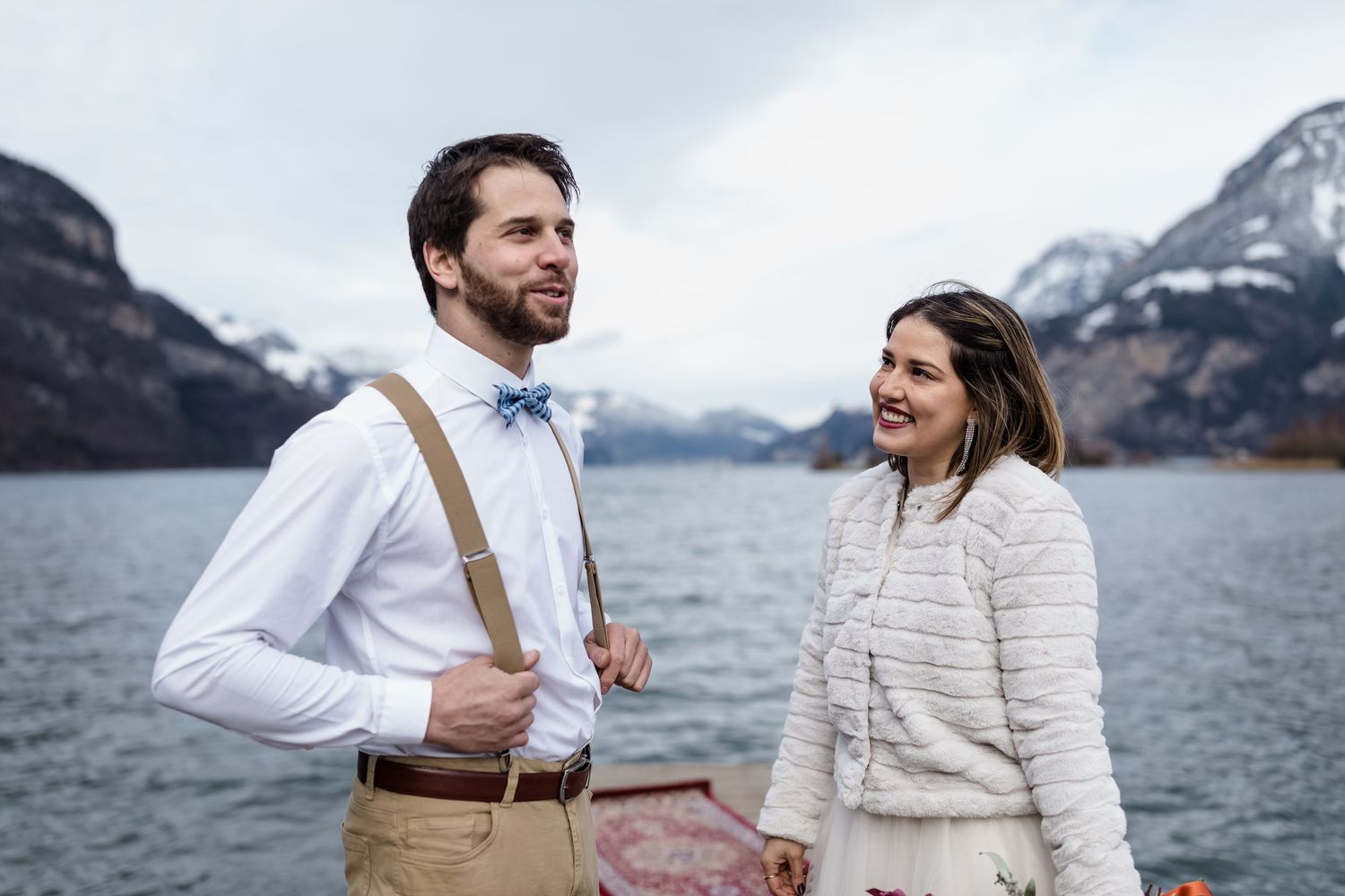 A couple in formal attire poses near a lake with snow-capped mountains in the background.