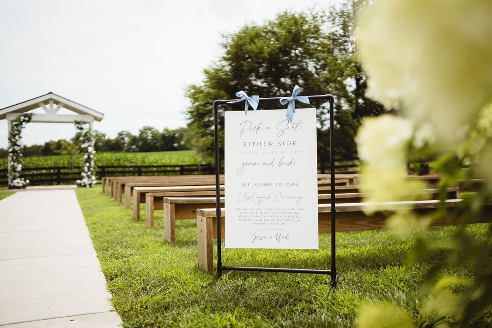 Wooden benches line a pathway leading to a wedding ceremony site with a welcome sign and floral arch in a garden setting.