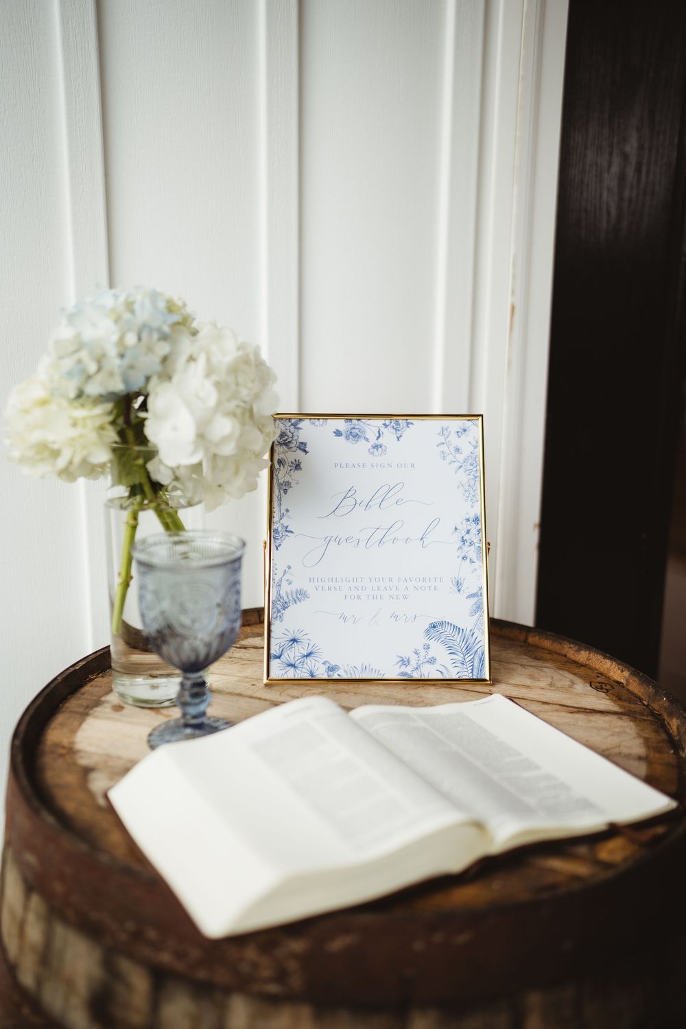 A rustic wooden plank displays an open white guestbook with elegant decorative cards and blue-tinted flower vases.