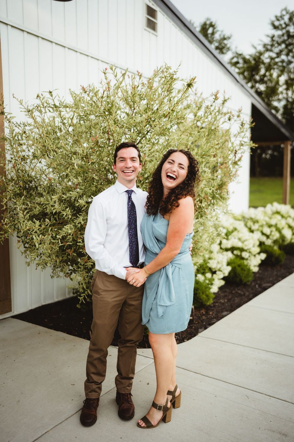 A couple shares a joyful moment together on a sidewalk in front of white shrubbery and landscaping.