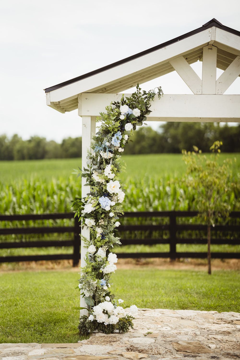 Elegant floral wedding arch with white flowers decorates a rustic farm setting with wooden fences and green pastures.